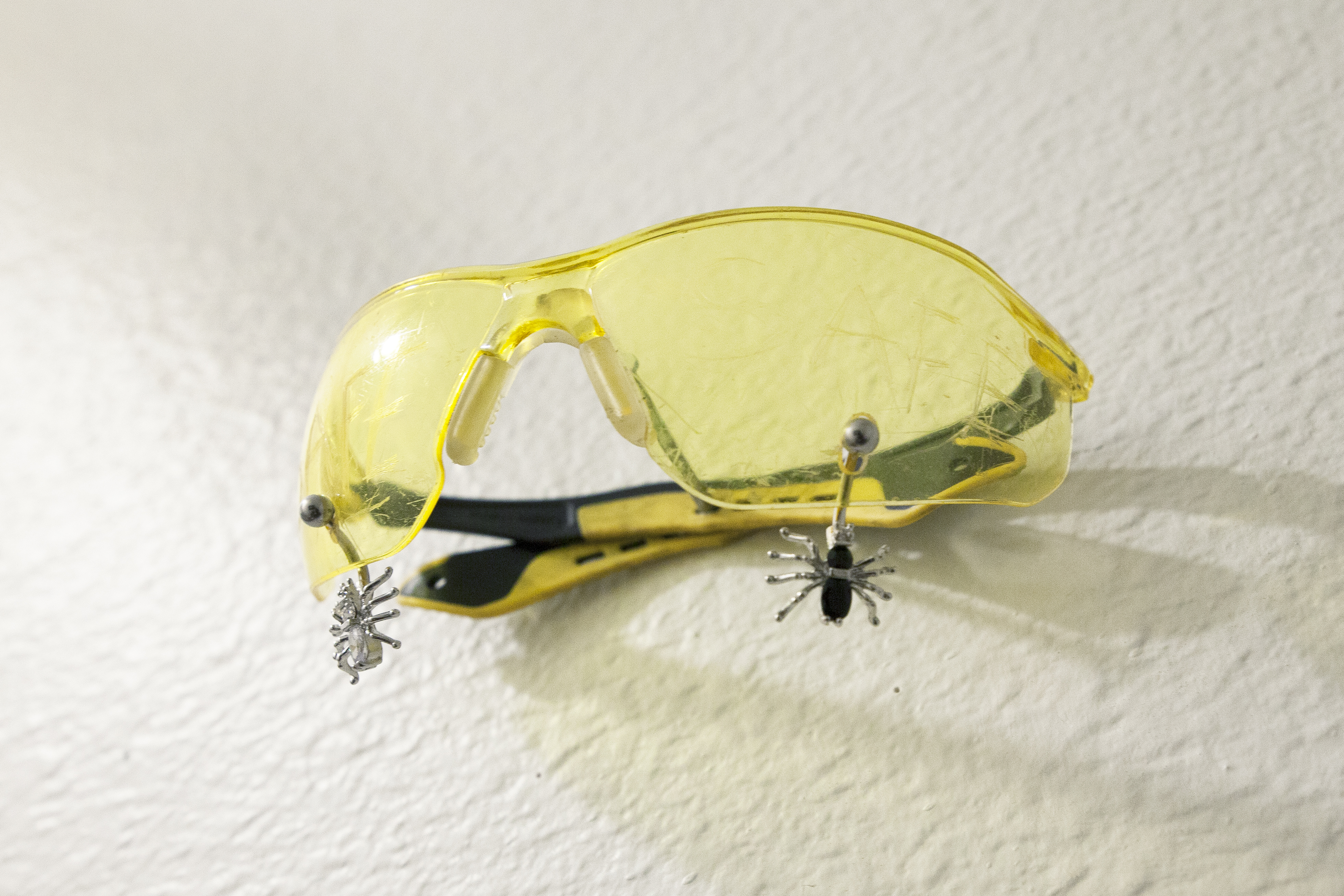 Yellow safety goggles with spider belly piercings through them hung on a white wall